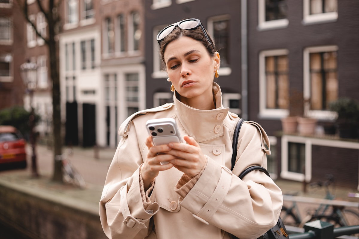 A young woman in a beige trench coat looks down at her mobile phone with a sad, weary, and disappointed expression while standing outdoors in an urban setting.